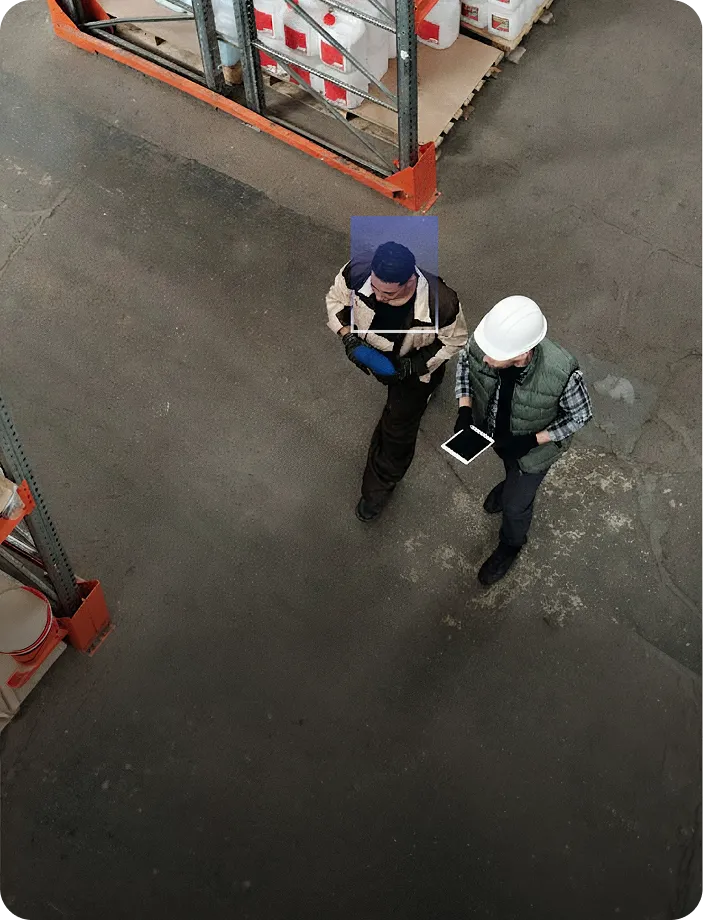 Two warehouse workers walking side by side on a warehouse floor, one holding a tablet and wearing a white hard hat.