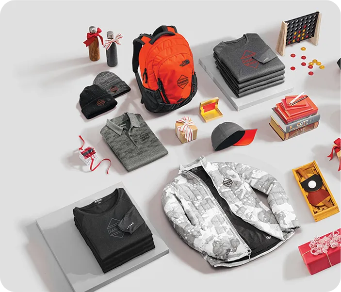 Collection of outdoor gear including an orange and black backpack, gray polo shirt, black folded shirts with logos, a white and gray camouflage jacket, beanies, a red and black cap, and small gift boxes arranged on a light surface.