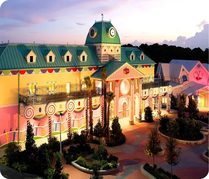 Colorful whimsical building with a green roof, candy-themed decorations, palm trees, and warm lighting at sunset.