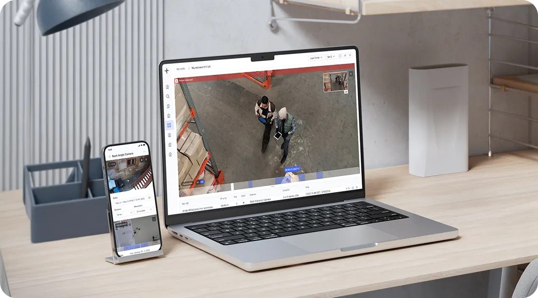 Laptop and smartphone on a wooden desk displaying security camera footage of people in a warehouse.