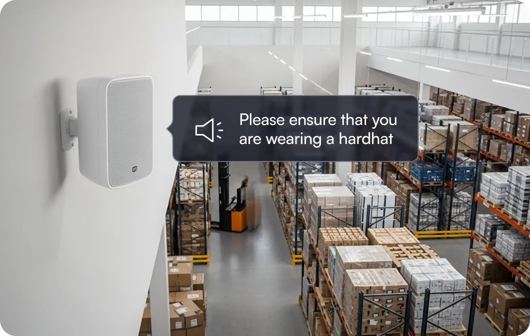 Warehouse interior with stacked pallets and a white speaker mounted on a wall displaying a message: 'Please ensure that you are wearing a hardhat.'