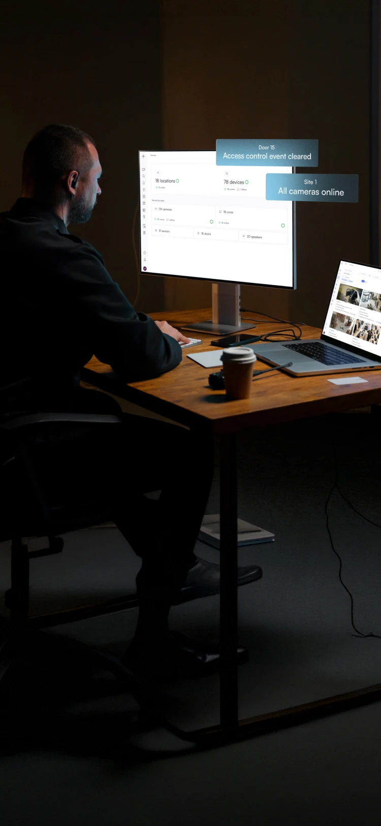 Man sitting at a desk in a dark room, monitoring security system statuses on a large computer screen and a laptop with surveillance camera feeds.