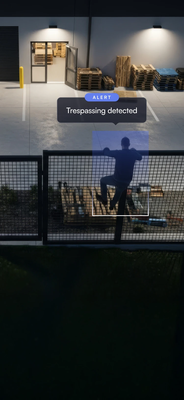 Person climbing a metal fence at night near a warehouse with stacked pallets and an open door, with an alert message saying Trespassing detected.