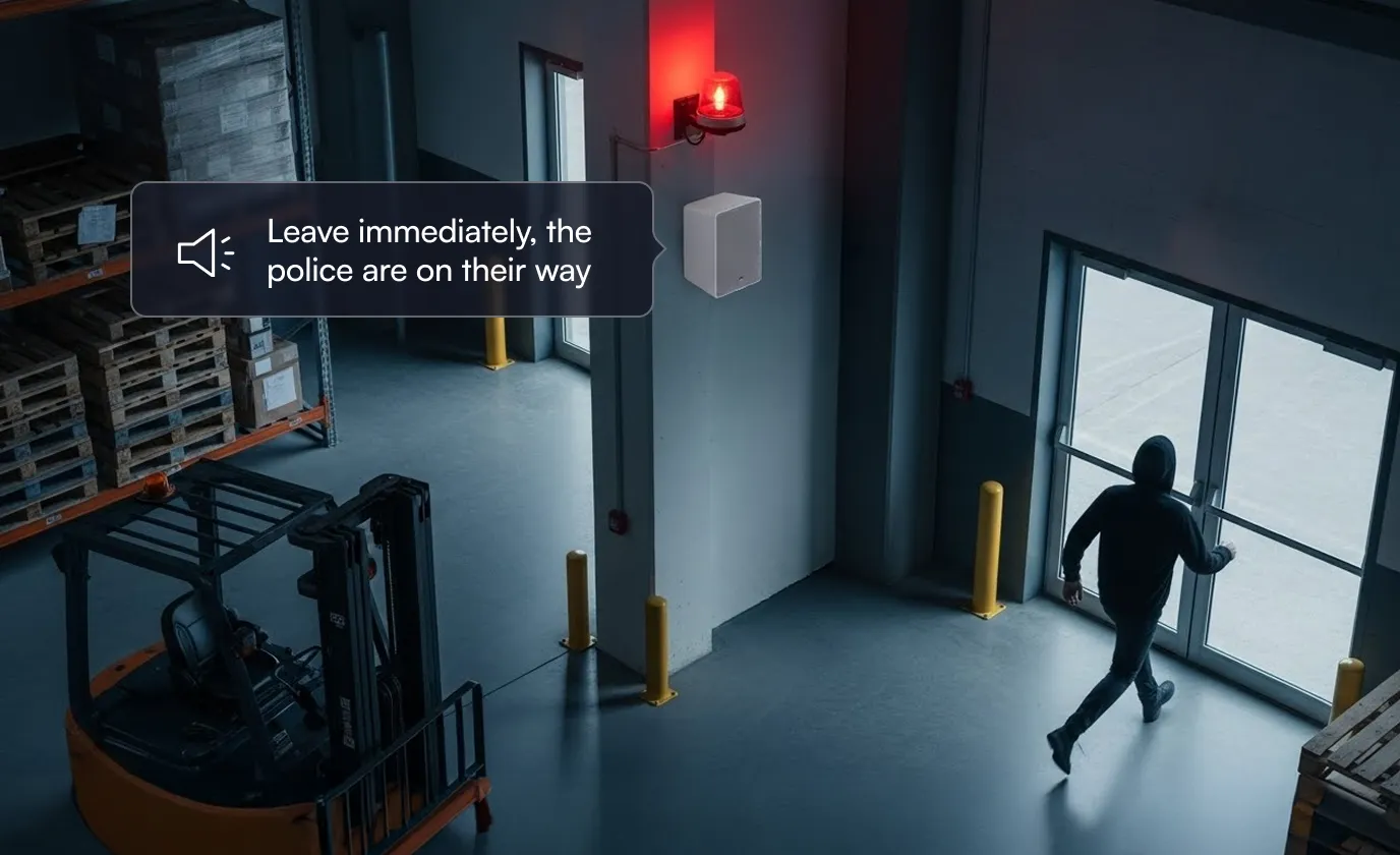 Person wearing a hood walking quickly towards an exit door in a warehouse with a security alarm flashing red and warning message saying police are on their way.