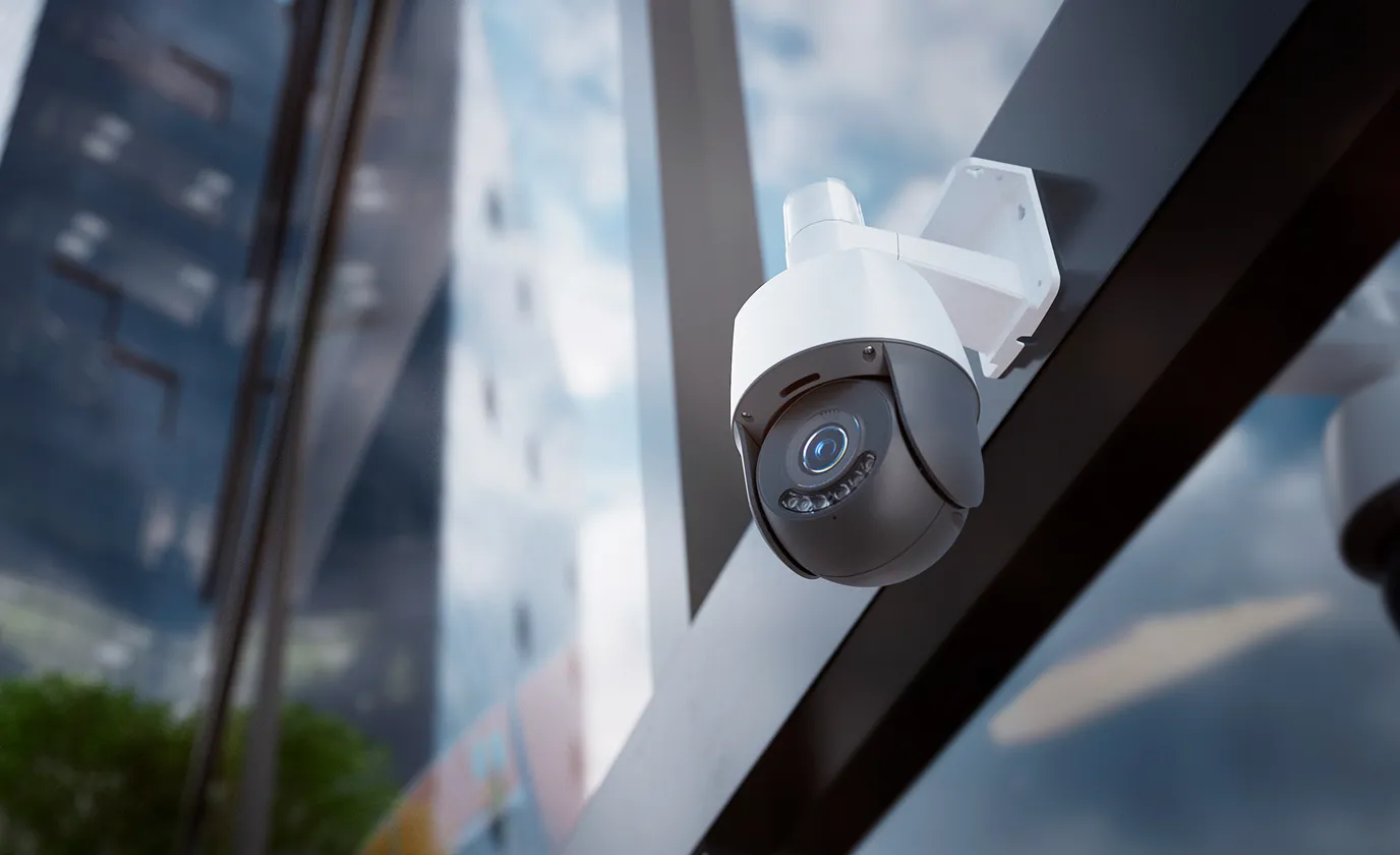 Modern outdoor security camera mounted on a building with glass windows reflecting the sky.