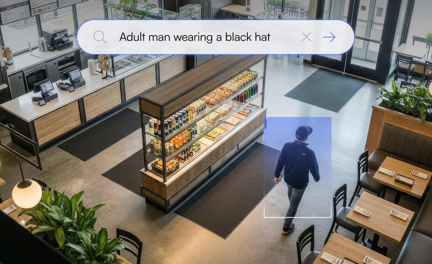 Overhead view of a man wearing a black hat and dark jacket walking inside a modern café near a display case with food and drinks.