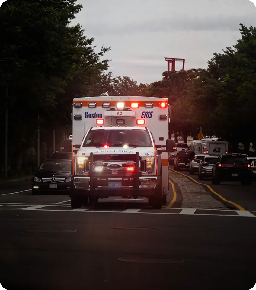 Boston EMS ambulance with flashing lights driving on a street with traffic and trees on both sides.