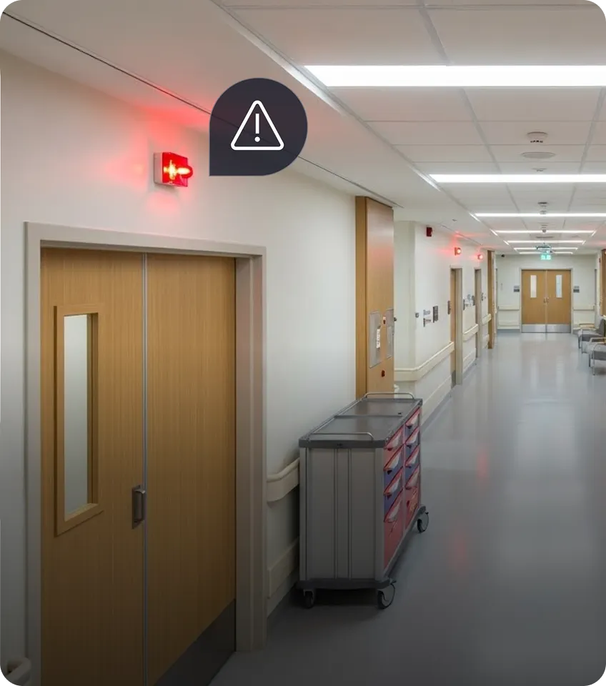 Hospital corridor with closed wooden double doors under a red alarm light and a medical supply cart nearby.