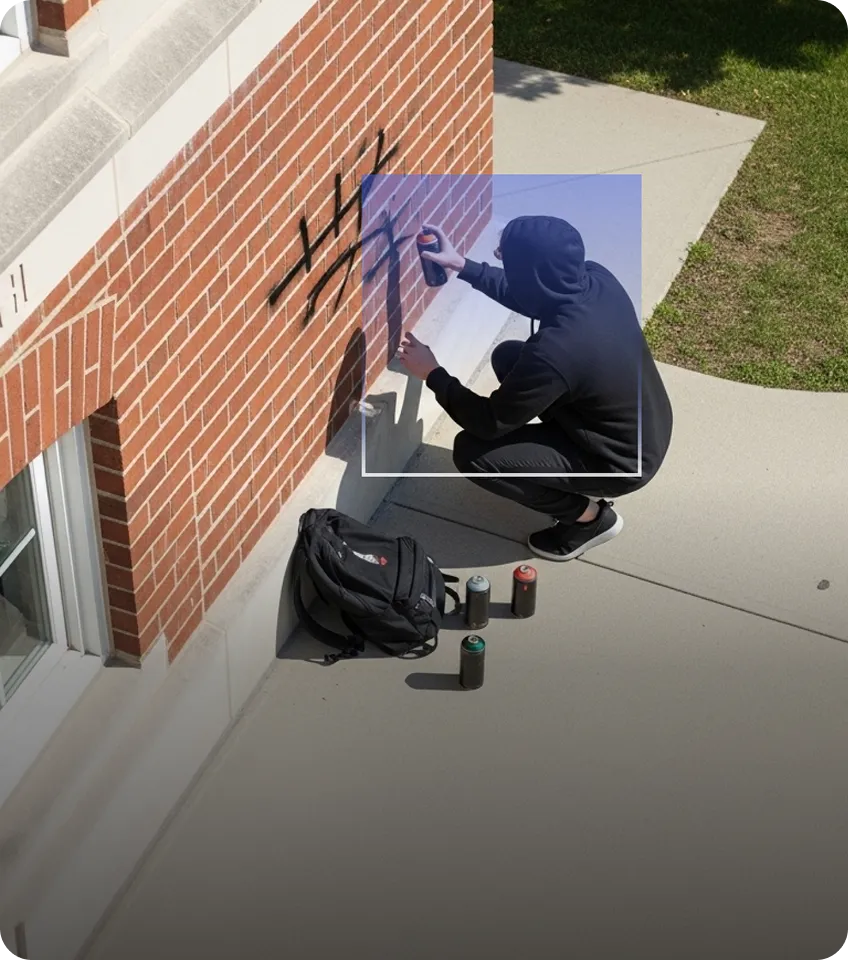 Person in black hoodie crouching while spray painting graffiti on a brick wall near a backpack and three spray paint cans on the ground.