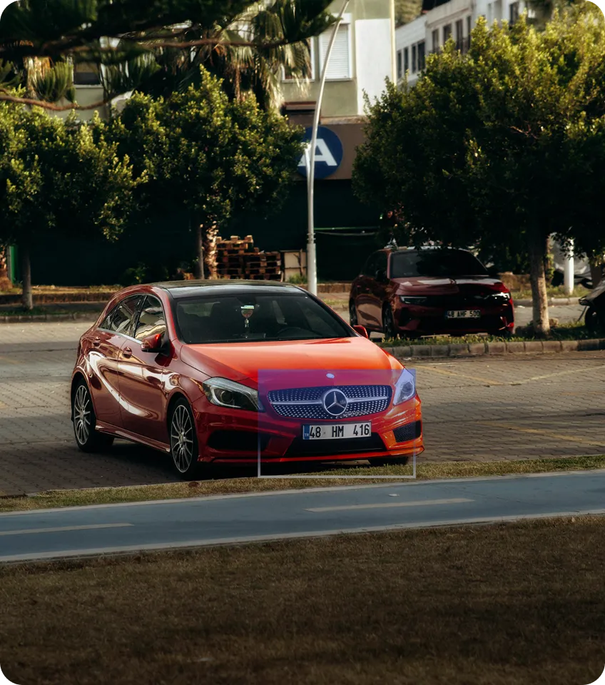 Red Mercedes-Benz hatchback parked on a paved area near green trees and another red car in the background.