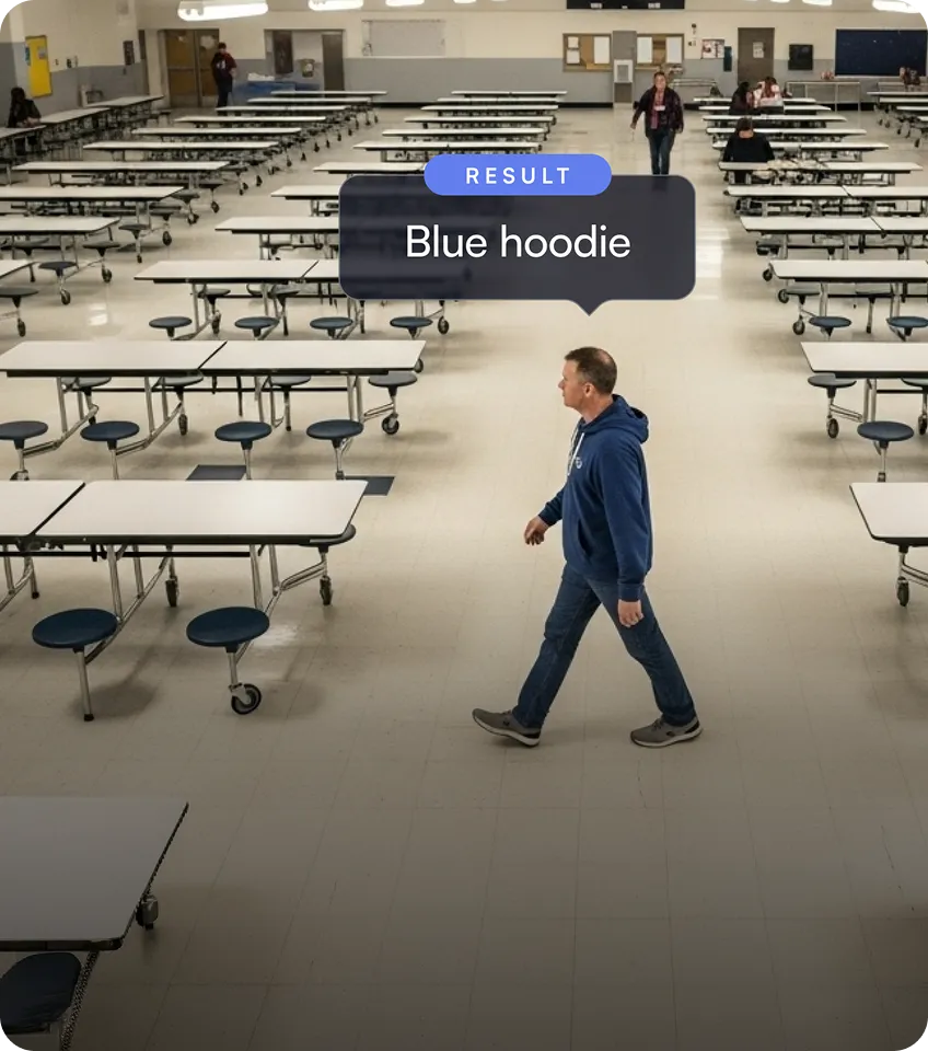 Man wearing a blue hoodie walking through an empty cafeteria with rows of tables and stools.