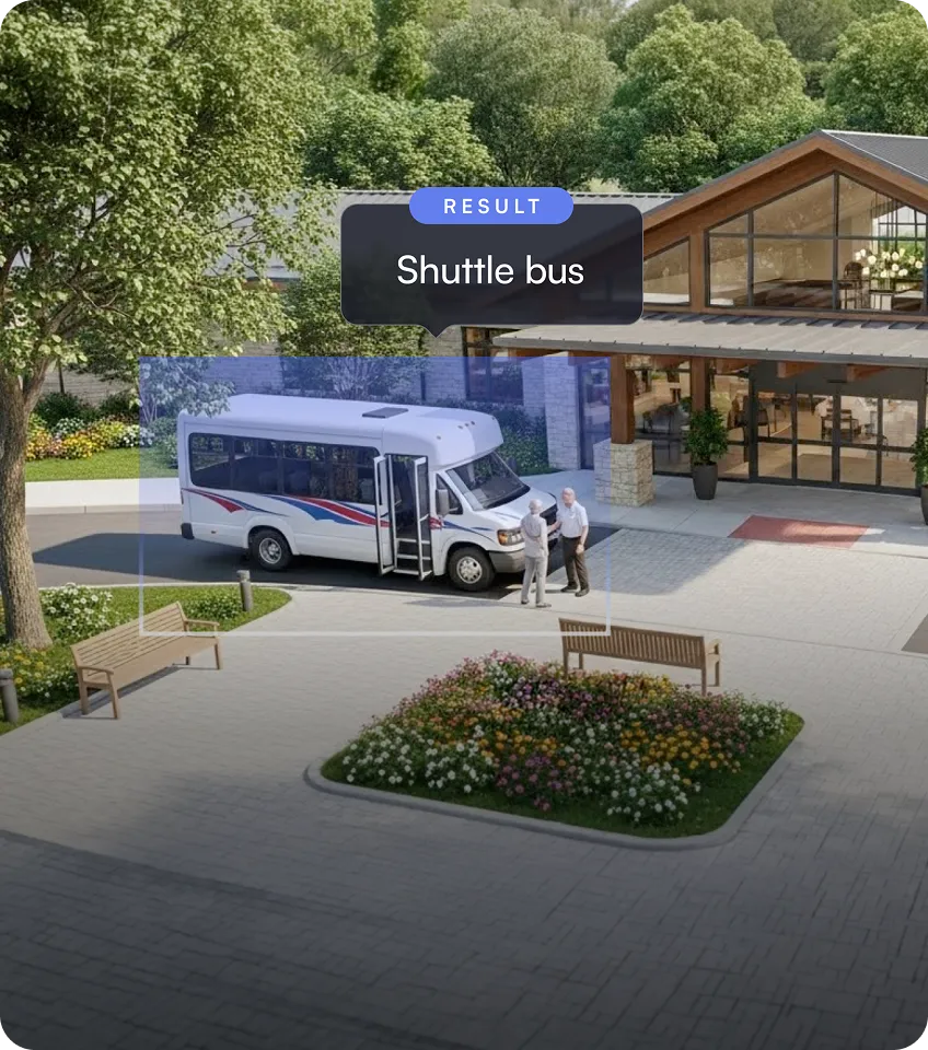White shuttle bus with red and blue stripes parked outside a building entrance with two people standing nearby.