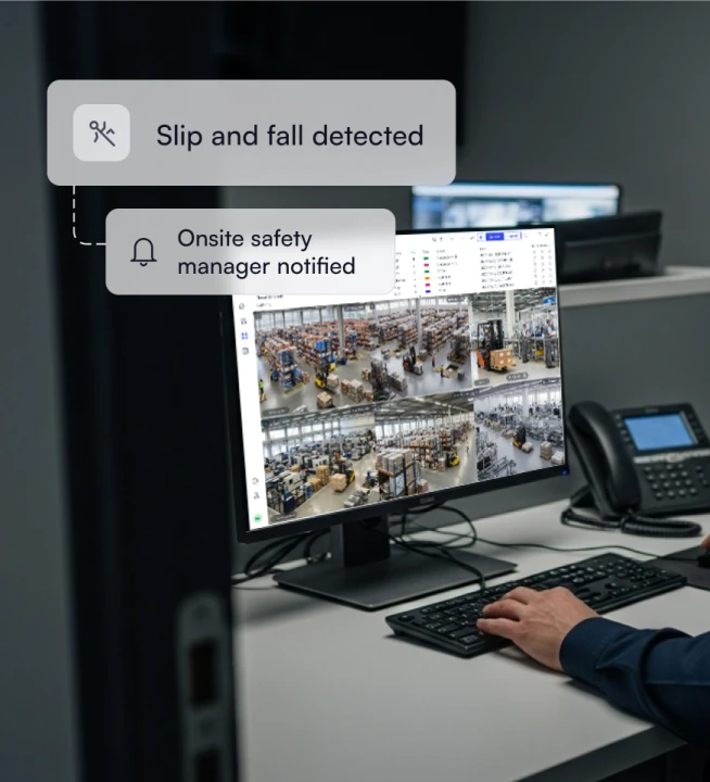 Computer monitor displaying warehouse surveillance video feeds showing equipment and storage racks, with notifications stating 'Slip and fall detected' and 'Onsite safety manager notified.'