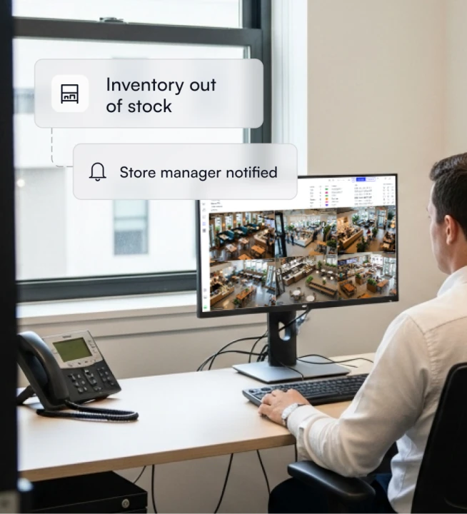 Person sitting at a desk monitoring multiple store security camera feeds on a computer, with notifications showing 'Inventory out of stock' and 'Store manager notified'.