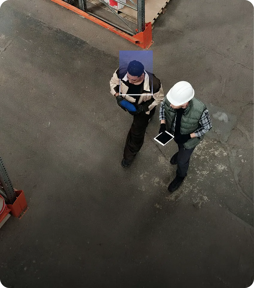 Two workers walking inside a warehouse, one holding a blue clipboard and the other wearing a white hard hat and holding a tablet.
