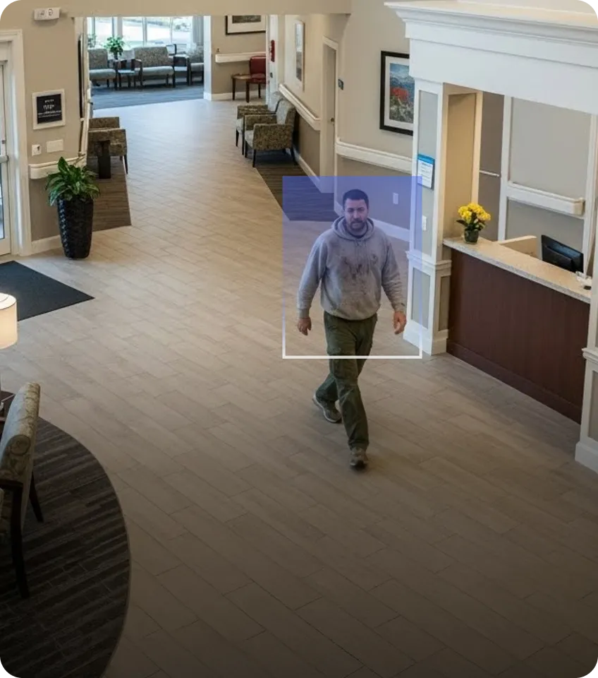 Man in a gray hoodie walking through a clean, modern lobby with beige tile flooring and a reception desk with yellow flowers.