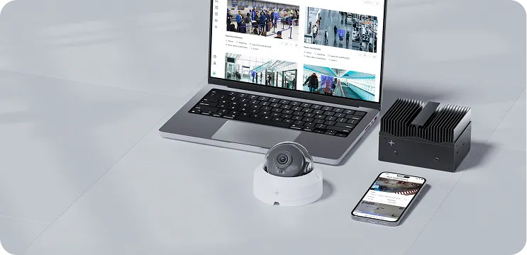 Laptop displaying security camera footage, a dome security camera on a white base, a black heat sink device, and a smartphone showing a security app on a white table.