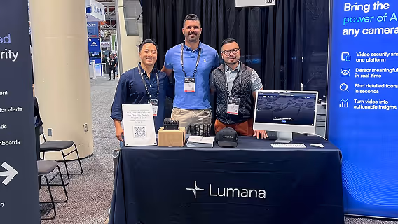Three men standing behind a Lumana booth with a computer and promotional materials at a trade show.