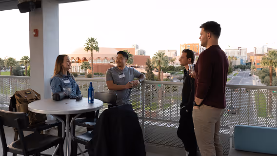 Four people casually talking on an outdoor balcony overlooking a city street with palm trees and buildings.