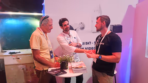 Three men with conference badges around a small high table, discussing papers and brochures indoors.