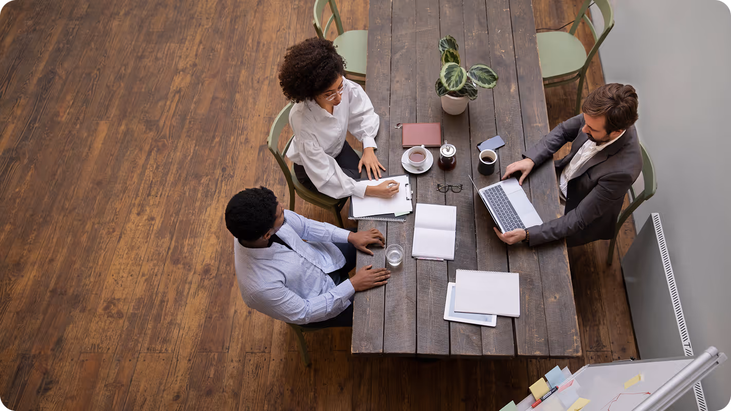 Three colleagues sitting around a wooden table with notebooks, laptop, coffee, and a plant, engaged in discussion.