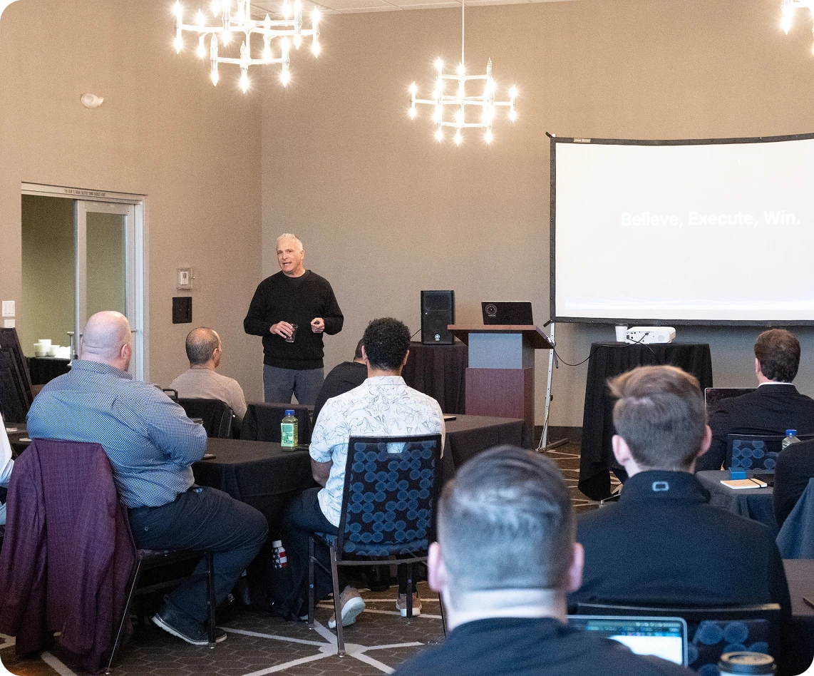 Speaker presenting to an audience seated at tables in a conference room with a projector screen displaying the text 'Believe, Execute, Win.'