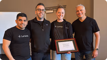 Four people standing indoors; one woman in the center holding a framed award certificate.