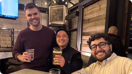 Three men smiling and holding drinks while seated at a table in a warmly lit indoor setting.