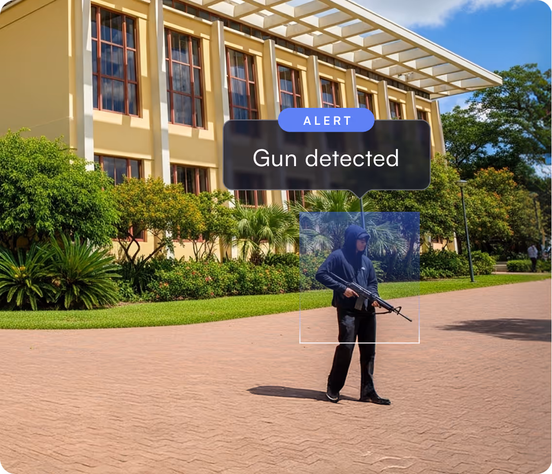 Person in black hoodie holding a rifle walking outside a building with an alert message reading 'Gun detected'.