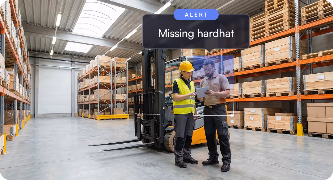 Warehouse interior with two workers near a forklift; one worker wears a yellow hardhat and vest, and an alert above them says 'Missing hardhat'.