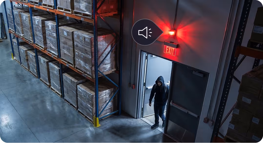 Man wearing a hoodie entering a warehouse through an emergency exit door under a red EXIT sign.