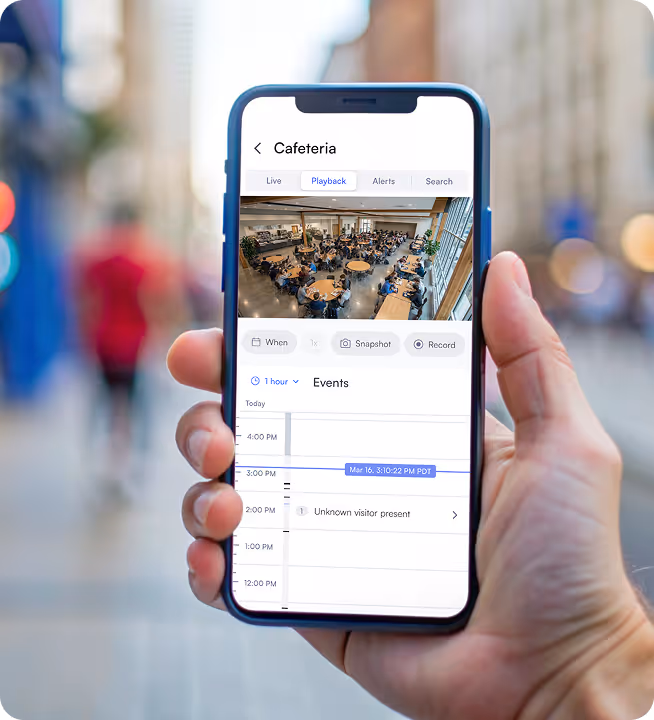 Hand holding smartphone showing cafeteria surveillance camera interface with playback and event timeline indicating unknown visitor presence.