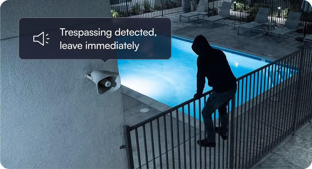 Person wearing a hoodie climbing over a fence near a lit swimming pool with a speaker displaying the message 'Trespassing detected, leave immediately.'
