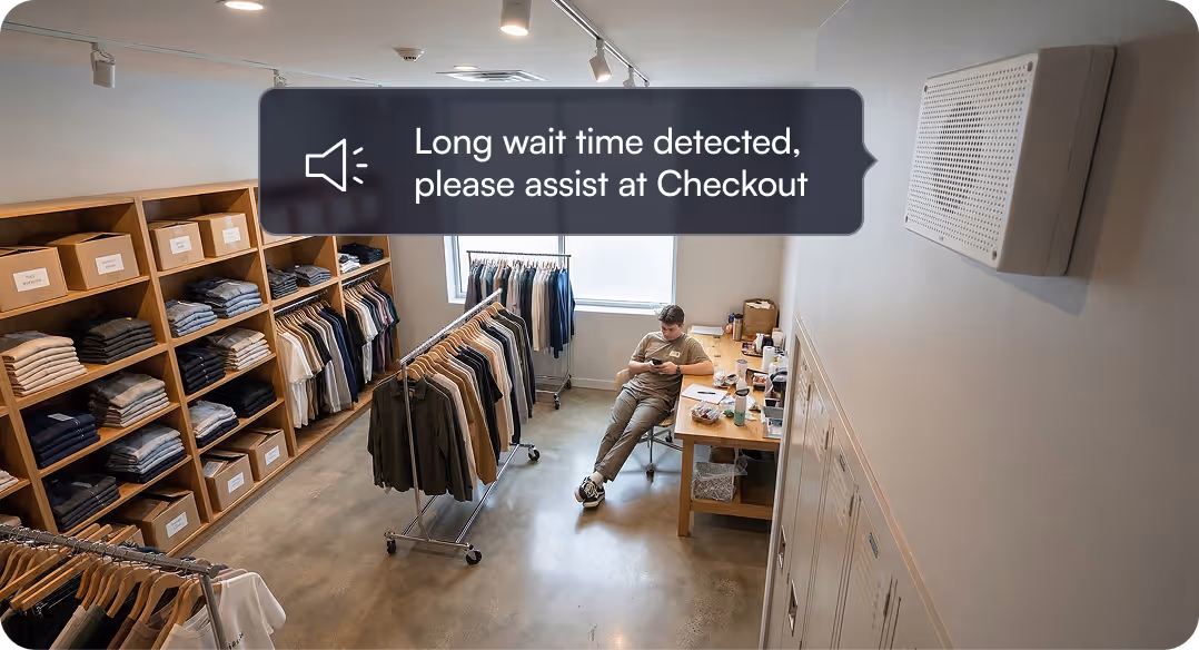 Retail employee sitting at a desk in a clothing store with racks of clothes and a notification saying 'Long wait time detected, please assist at Checkout.'