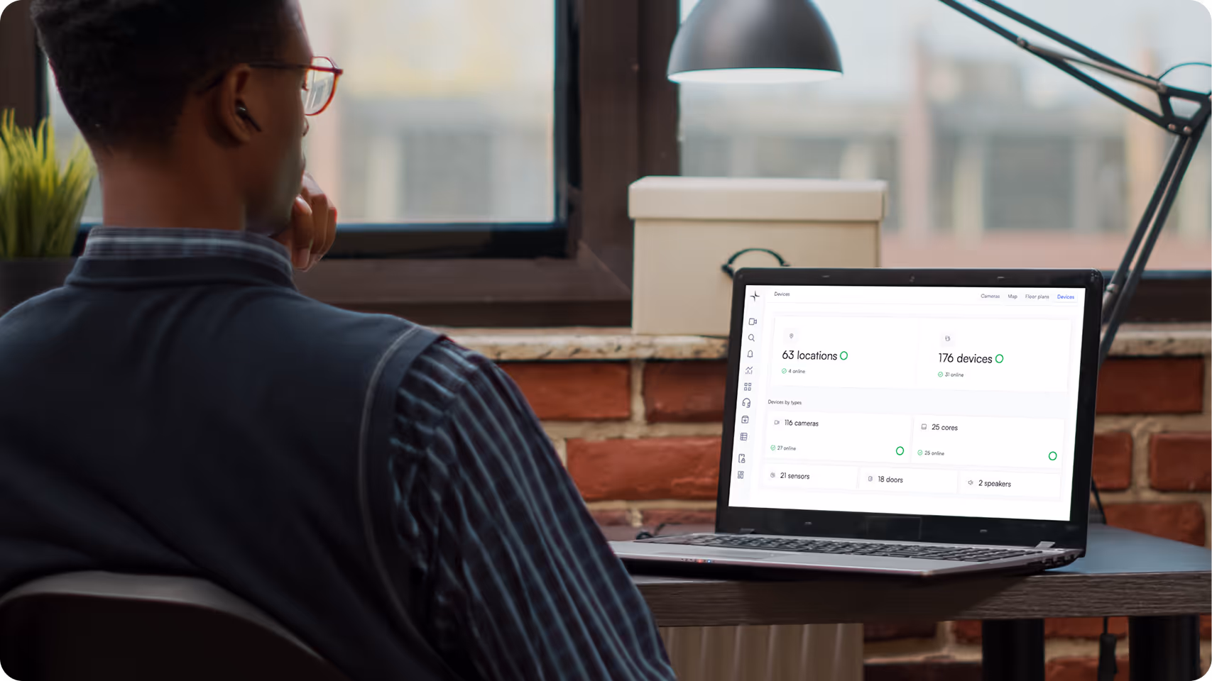 Person sitting at a desk looking at a laptop screen displaying a dashboard with device statistics including locations, devices, cameras, cores, sensors, doors, and speakers.