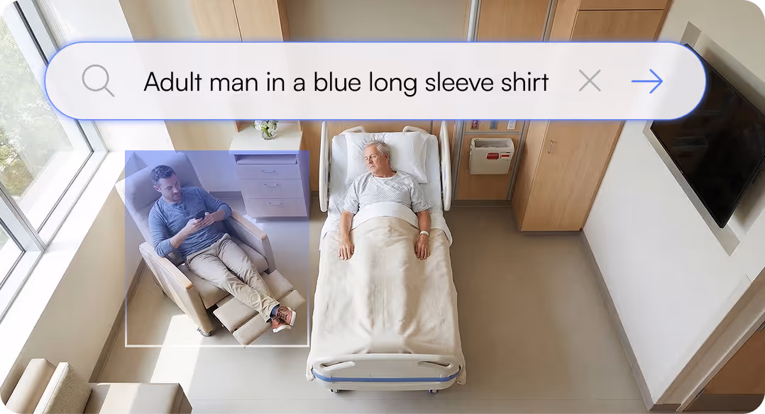 Hospital room with an elderly man lying in bed and a younger man in a blue long sleeve shirt sitting on a recliner chair using a smartphone.