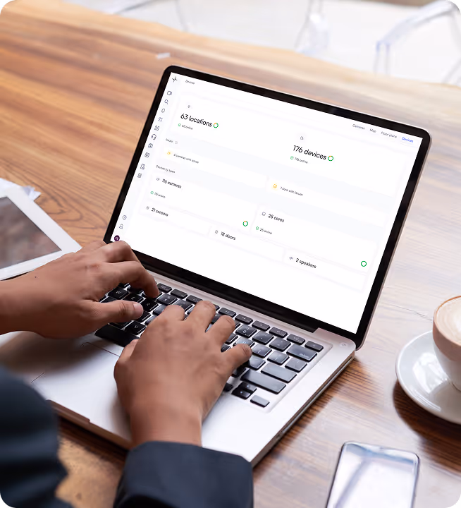Person typing on a laptop displaying a device and location management dashboard with counts of cameras, sensors, and speakers.