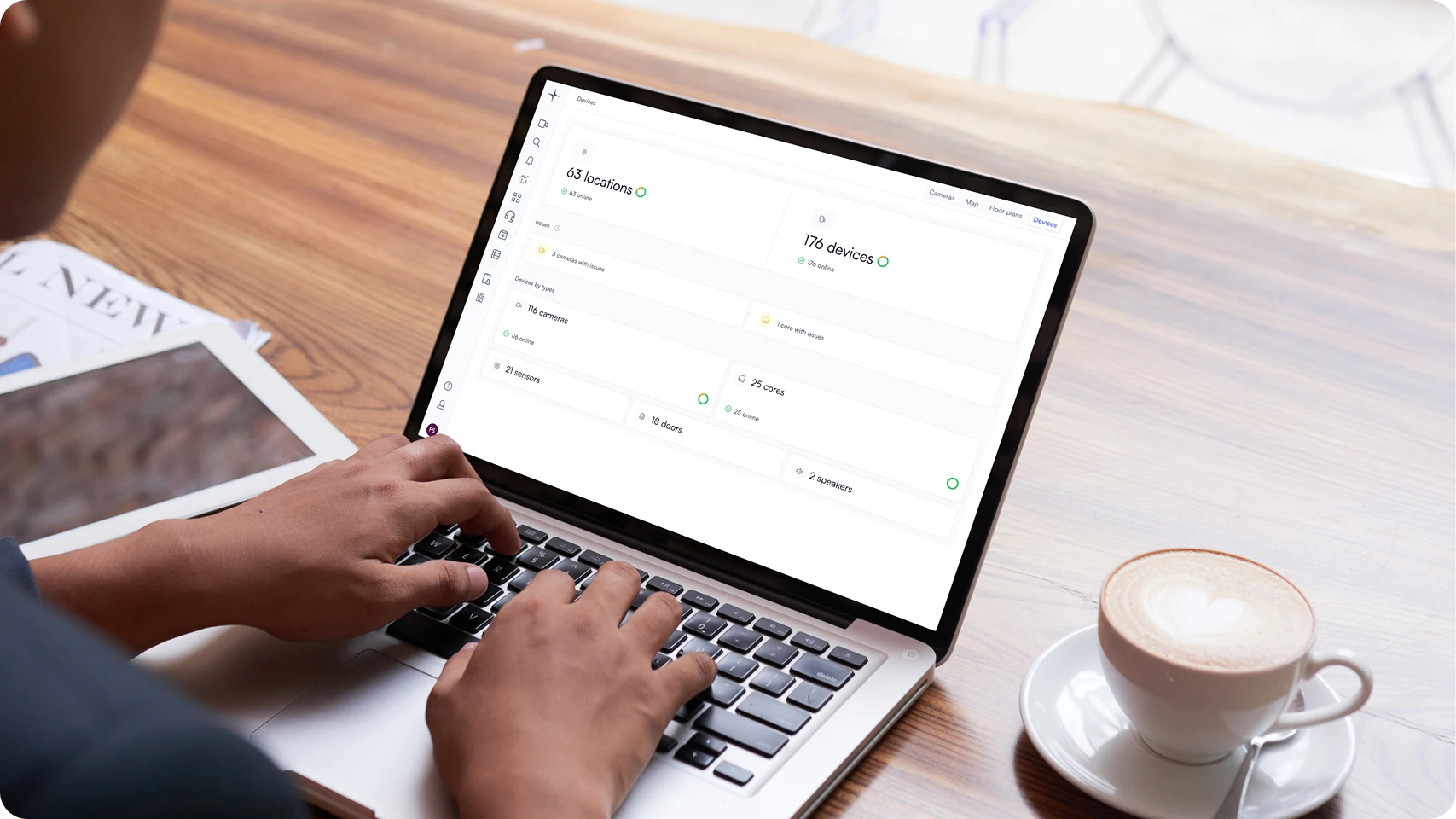 Person typing on a laptop showing a device monitoring dashboard, with a cup of coffee and a tablet on a wooden table.