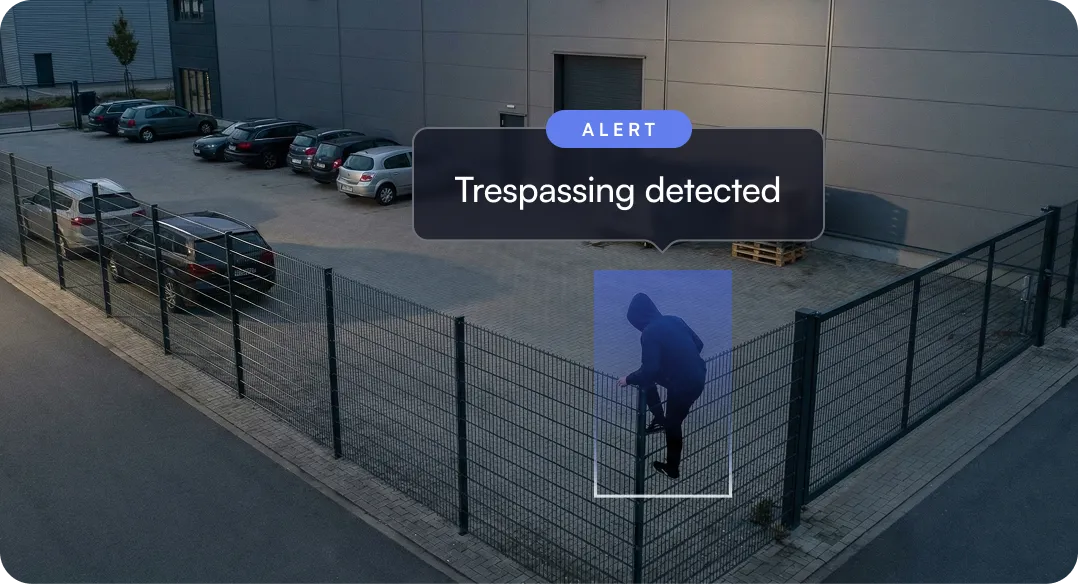 Person in a hoodie climbing over a fenced perimeter detected as trespassing near parked cars and a building.