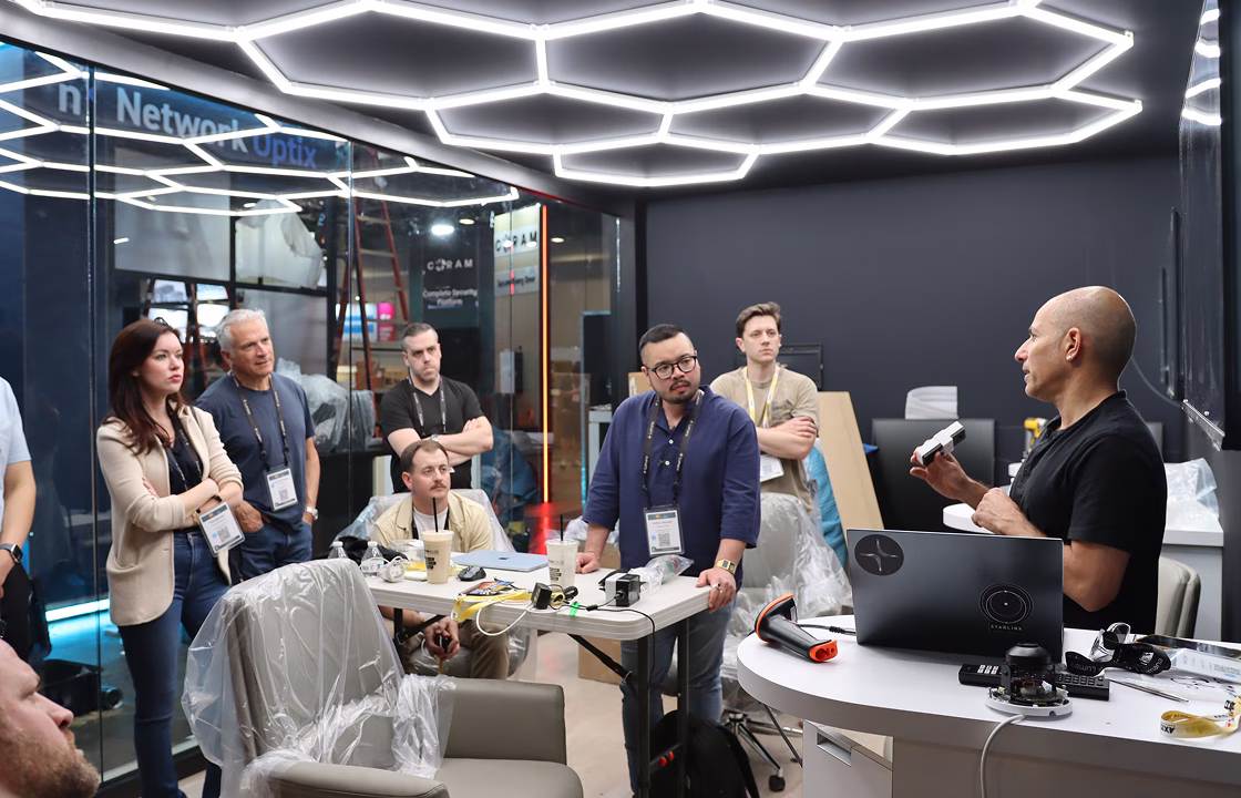 Group of people attentively listening to a man presenting with a device in a conference room with hexagonal ceiling lights.