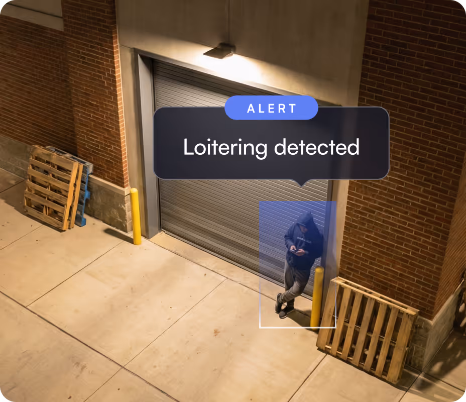 Security camera image showing a person in a hoodie leaning against a wall near a closed garage door with an alert box saying 'Loitering detected'.