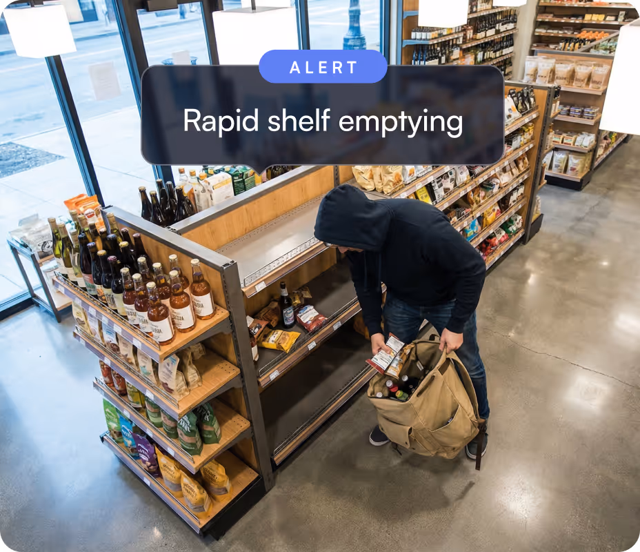 Man in a black hoodie stealing items from nearly empty store shelves into a large beige bag.
