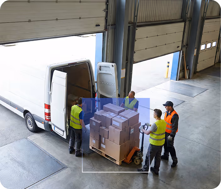 Four workers in safety vests loading cardboard boxes onto a pallet jack next to a white delivery van inside a warehouse.