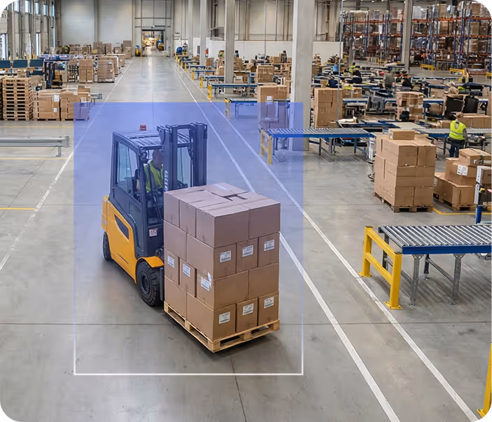Warehouse interior with a forklift carrying stacked cardboard boxes on a pallet, surrounded by shelves and workers handling packages.