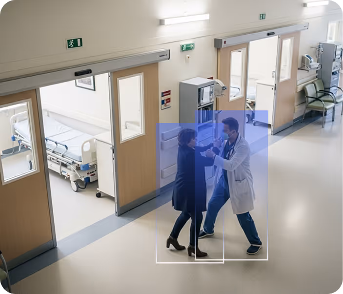 A doctor greeting or assisting a woman in a hospital corridor near patient rooms and medical equipment.