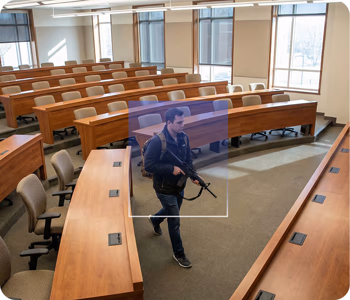 A young man carrying a rifle walks through a mostly empty university lecture hall with wooden curved desks and chairs.