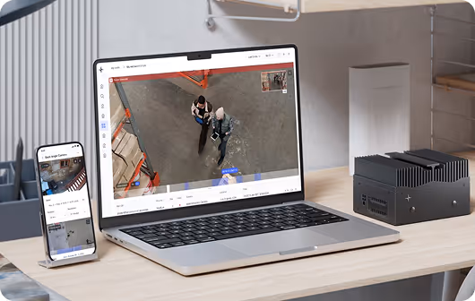 Laptop and smartphone on desk showing security camera footage of two people walking in a warehouse.