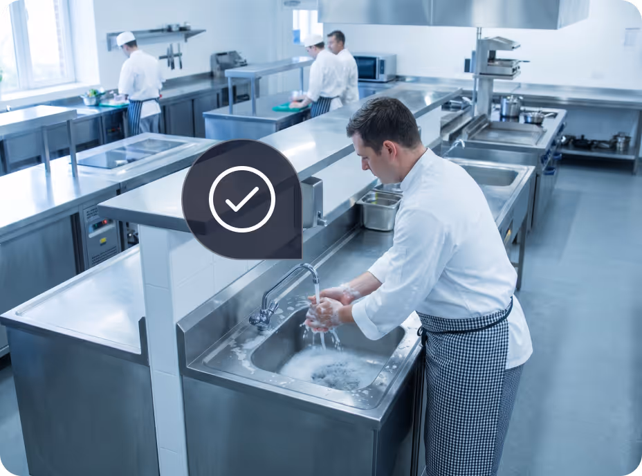 Chef washing hands with soap in a large stainless steel kitchen while other chefs work in background.