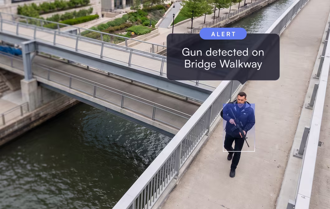 Man in blue jacket holding rifle walking on bridge walkway next to river with alert message above him.