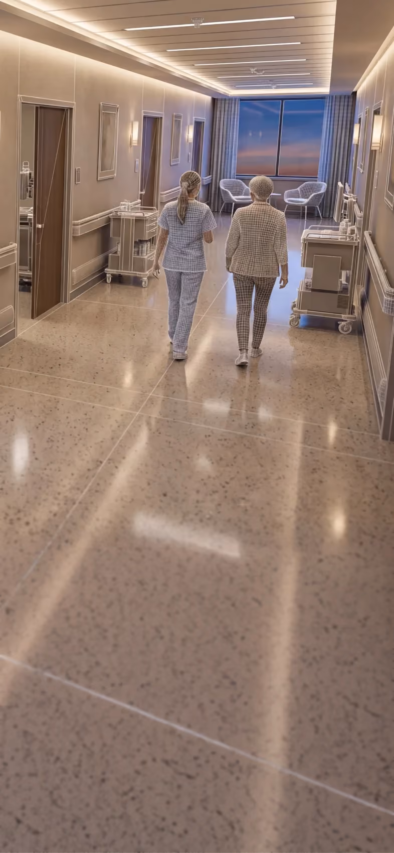 Two healthcare workers walking down a hospital corridor with carts and chairs near a window.